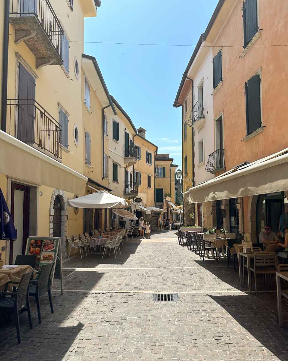 Blick in eine ruhige Altstadt-Gasse ohne Autos mit südlichem Flair an einem sonnigen Tag. Die Häuser sind bunt in Ockertönen und dreistöckig. Auf der Straße haben Cafés Sonnenschirme, Stühle und Tische aufgestellt. 