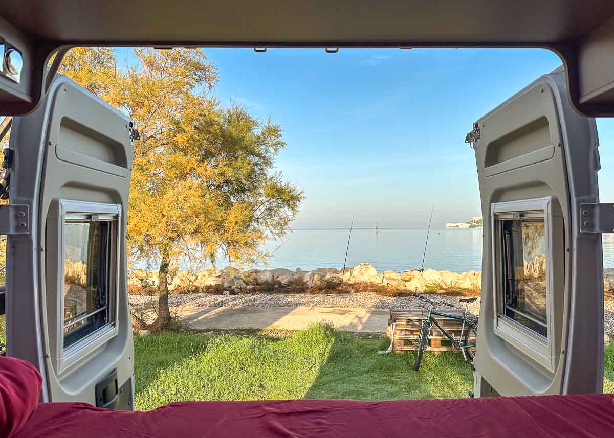 Blick durch die offenen Hecktüren eines Kastenwagen-Wohnmobils über ein rot bezogenes Bett auf das Meer. Auf der Wiese am Ufer steht ein Herbstbaum.