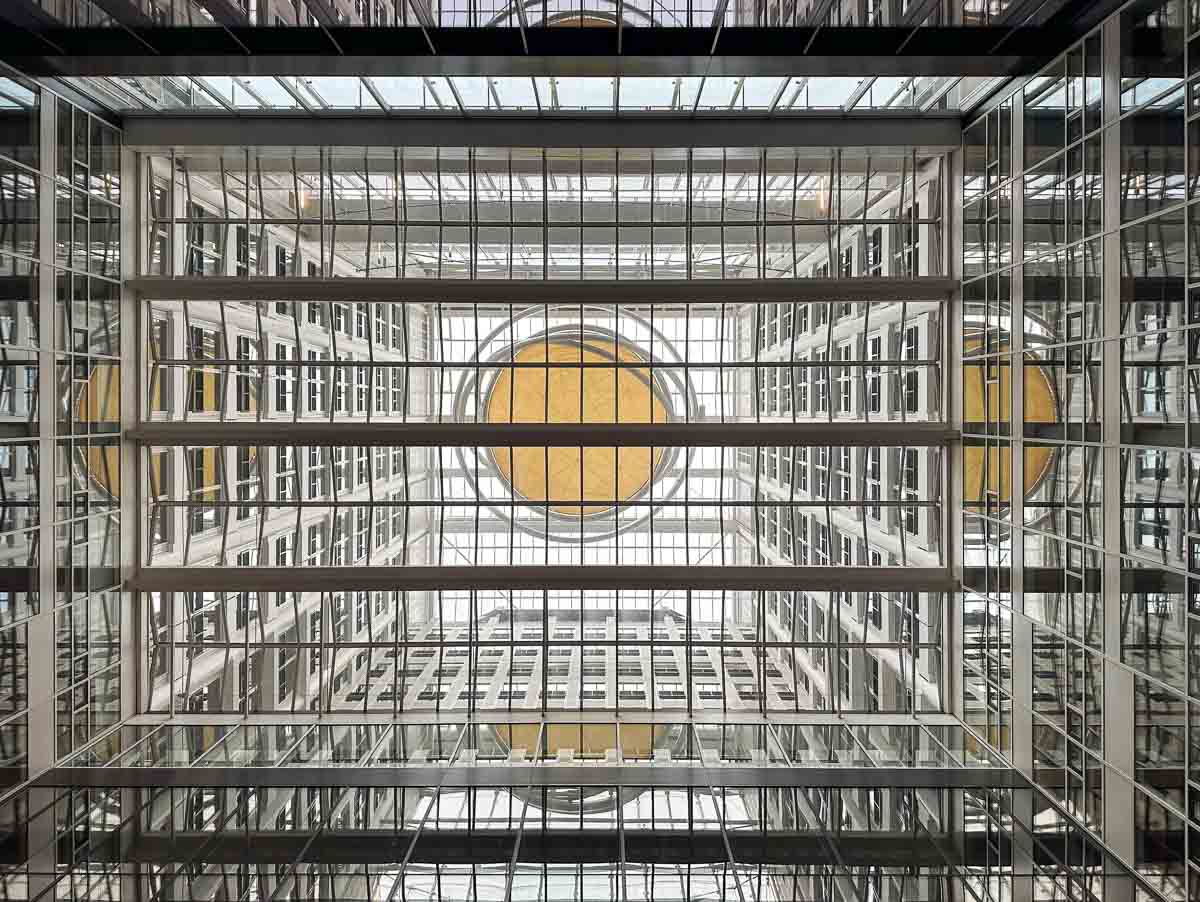 Blick nach oben im Hof eines Wolkenkratzers durch ein Glasdach mit Gittermuster. Oben ist ein gelber Kreis angebracht.
