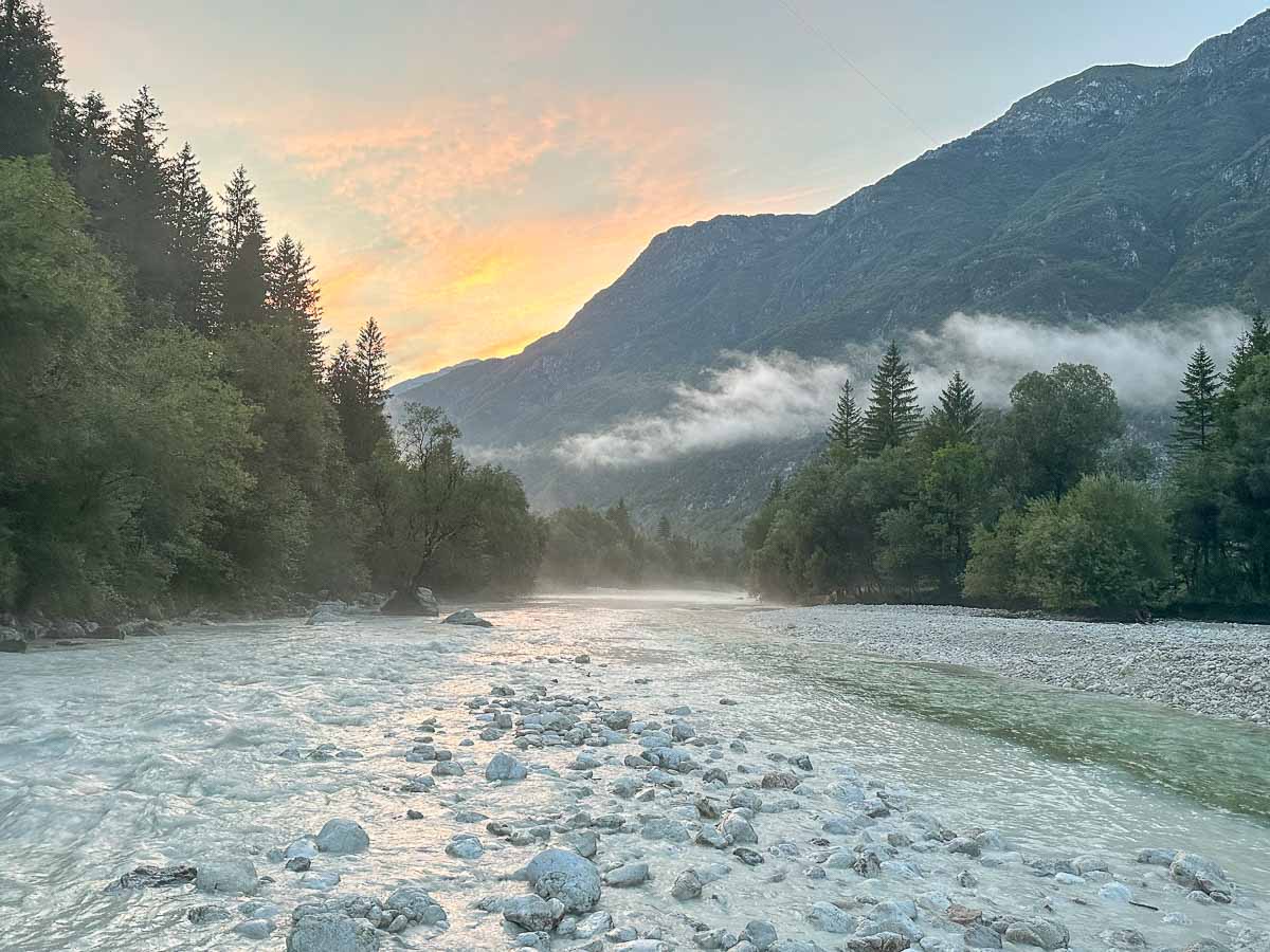 Abendstimmung an einer Flussmündung in den Bergen mit sprudelnd weißem Wasser und großen weißen Kieseln in den Flussbetten. Über dem Wasser liegt leichter Nebel. Am Ufer stehen Nadelbäume. Durch das Tal ziehen Wolkenfetzen.