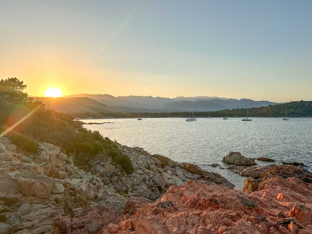 Sonnenuntergang hinter den Bergen über einer Meeresbucht. Im Vordergrund ist eine rote Felsenküste zu sehen.