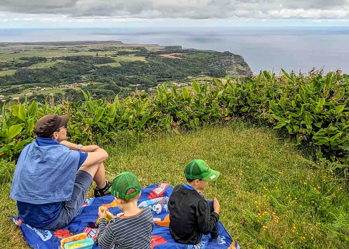 Auf einem Berg auf den Azoren genießen ein Vater und zwei Kinder die weite Aussicht über die atemberaubend wilde grüne Natur bis zum Atlantik bei einem Picknick.