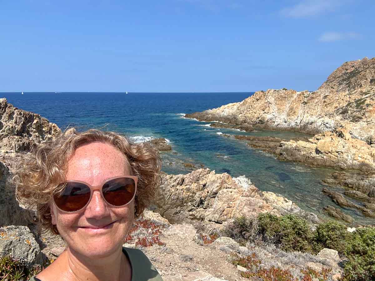 Eine Frau mit Sonnenbrille lacht freudestrahlend in die Kamera. Sie steht in einer atemberaubend schönen Küstenlandschaft mit zerklüfteten Felsen in einem türkisblauen Meer.