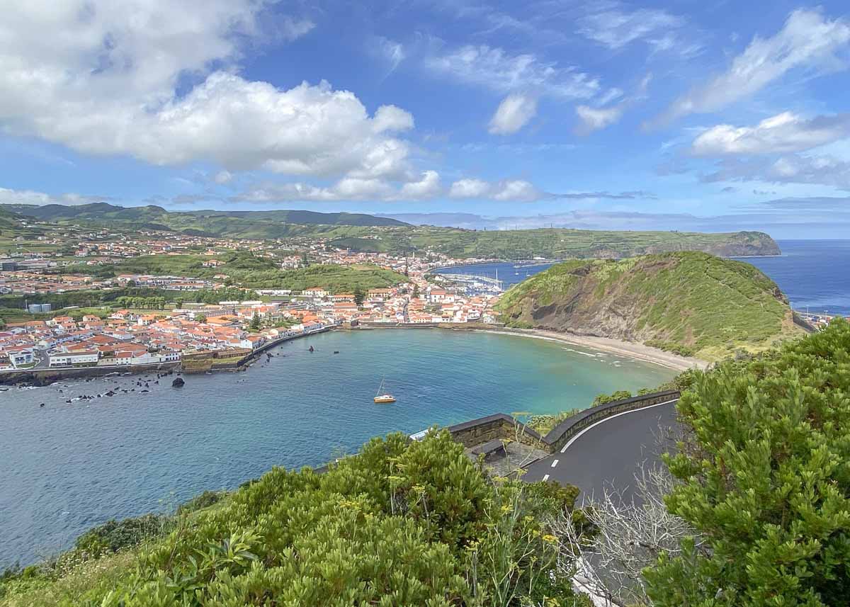 Blick von einem Berg über grüne Hügel auf die geschützte Hafenbucht der Insel Faial auf den Azoren, wo Häuser stehen und Schiffe im Wasser sind.