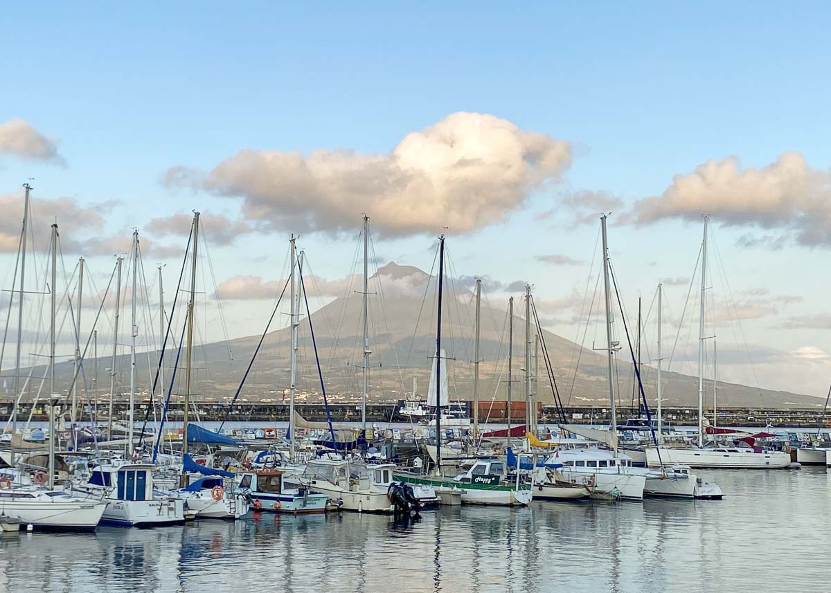 In einem idyllischen Yachthafen auf den Azoren schaukeln Segelboote am Abend. Im Hintergrund schient die Abendsonne auf einen Vulkan.
