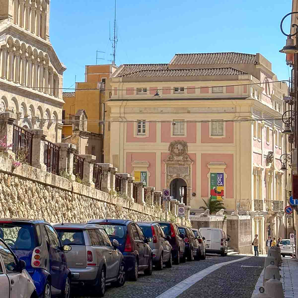 Eine Straße mit Parkplätzen läuft auf den prachtvollen Antico Palazzo della Citta in Cagiari Altstadt zu. Von oben links kommt ein Balustraden gesäumte zwei Straße. Rechts sieht man stilvolle, historische Straßenlaternen in der Altstadtstraße. 