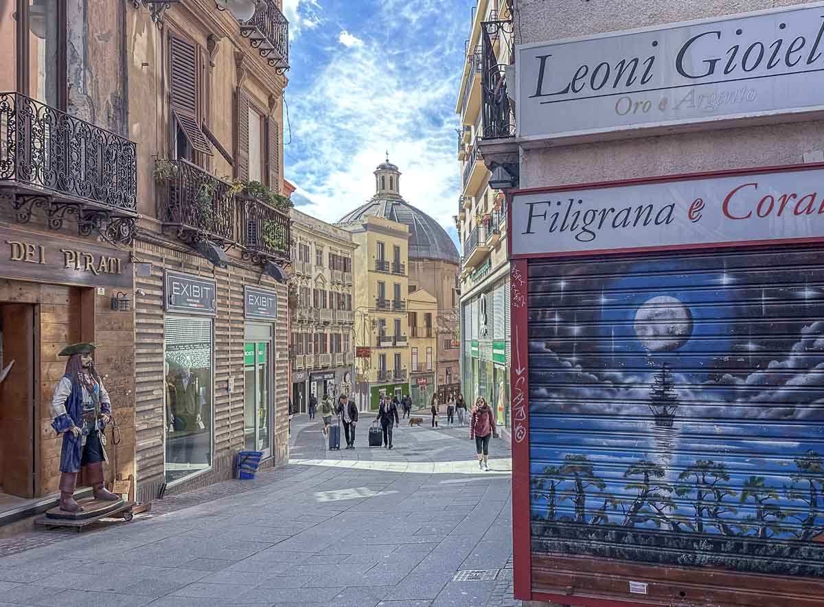 Blick in eine Fußgängerzone mit vielen Geschäften und einer Kuppel am Ende der Straße. Links steht eine Piratenfigur an einem Laden, rechts sieht man Street Art an einem geschlossenen Schaufensterrollo eines Juweliers. 