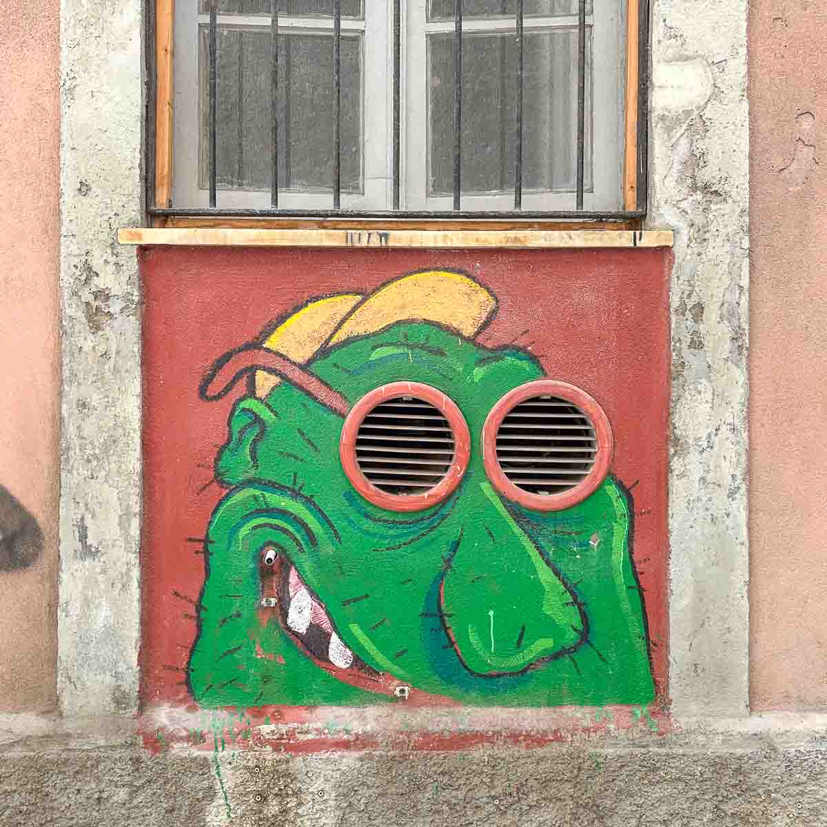 Street Art, die 2 Lüftungsschächte als Sonnenbrillengläser für ein grünes Comic-Gesicht nutzt.