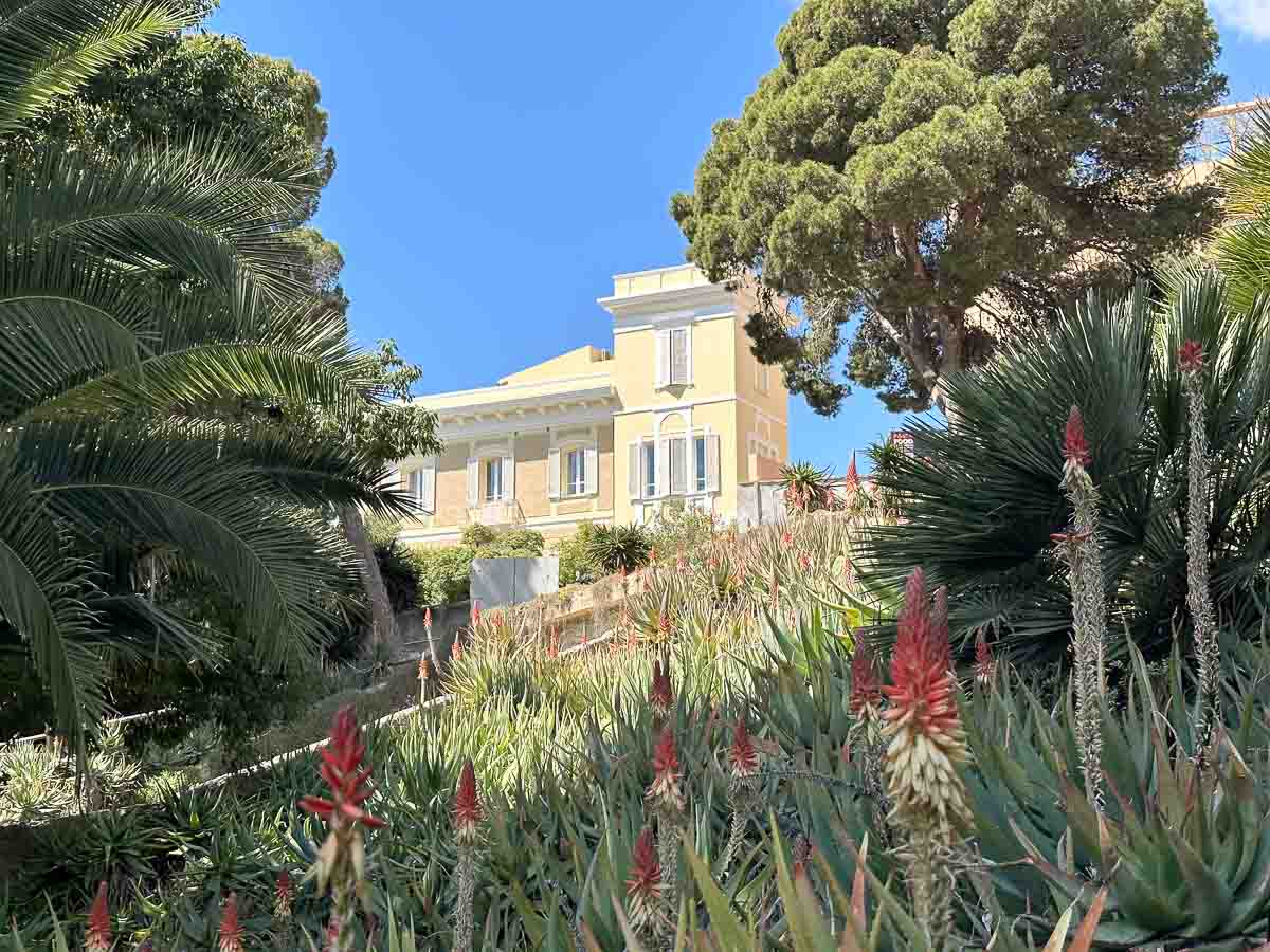 Eine Italienische Villa mit gelber Fassade trohnt zwischen Palmen und anderen Bäumen über blühenden Blumenbeeten im Botanischen Garten Cagliari auf Sardinien.