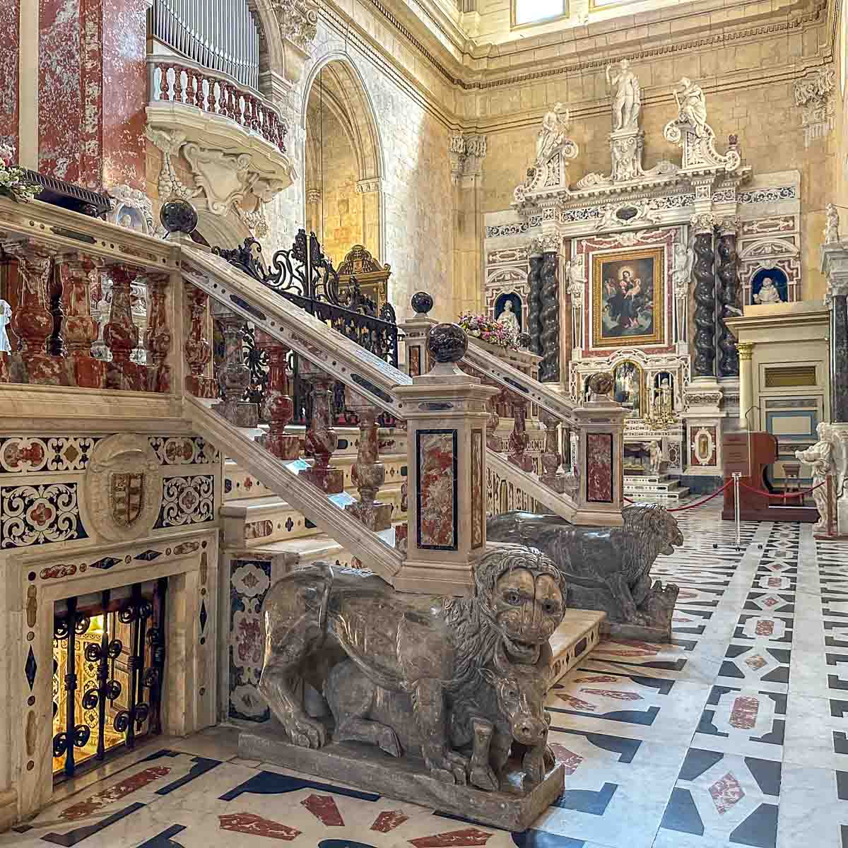Prachtvolles Inneres der Kathedrale von Cagliari mit Barockschmuck, unter anderem sieht man eine Treppenbalustrade mit Marmorlöwen, einen gemusterten Marmorboden und zwei Marmoraltäre mit viel Goldschmuck.