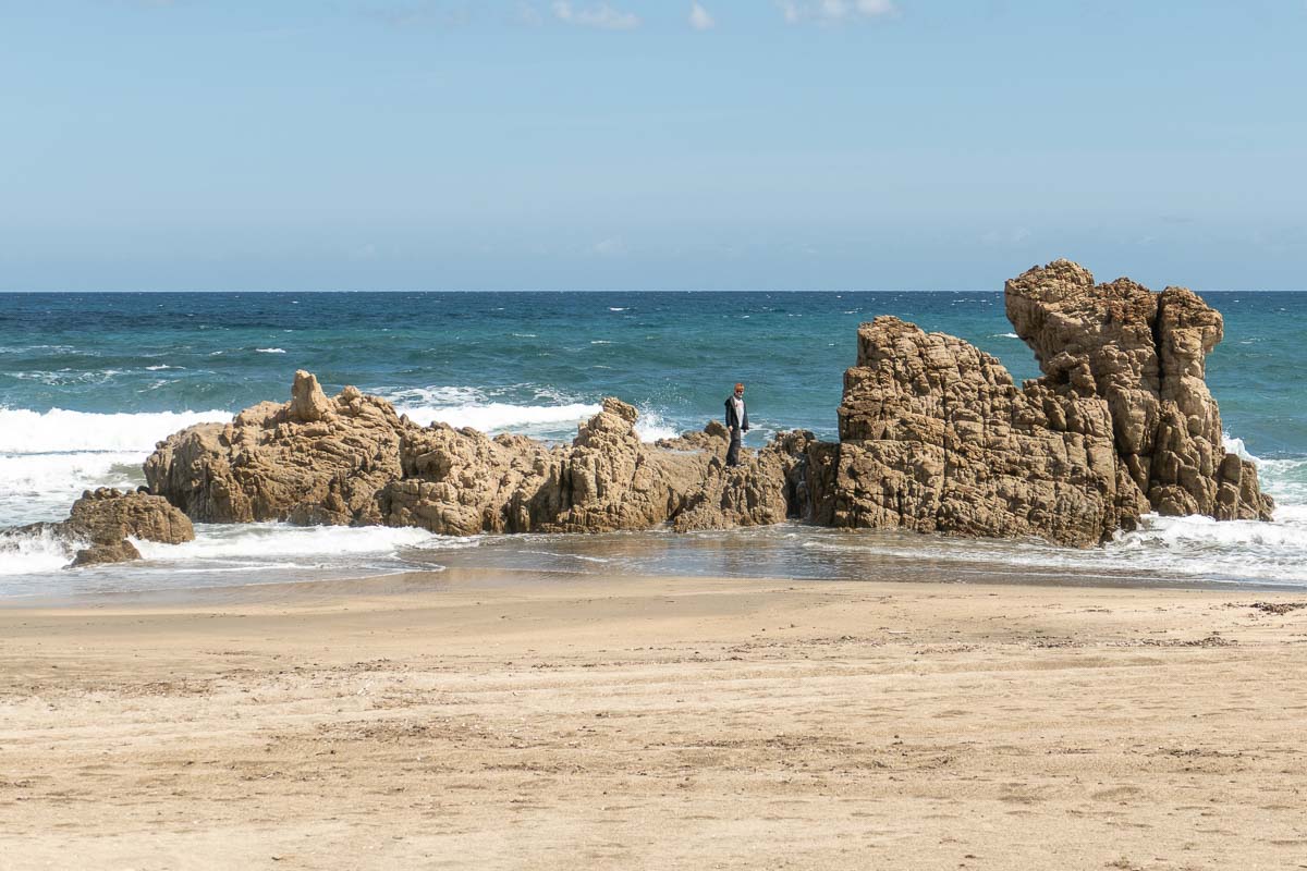 Ein großes Kind in Wanderschuhen klettert auf einem Felsenschiff in den flachen Brandungswellen eines Sandstrandes auf Sardinien