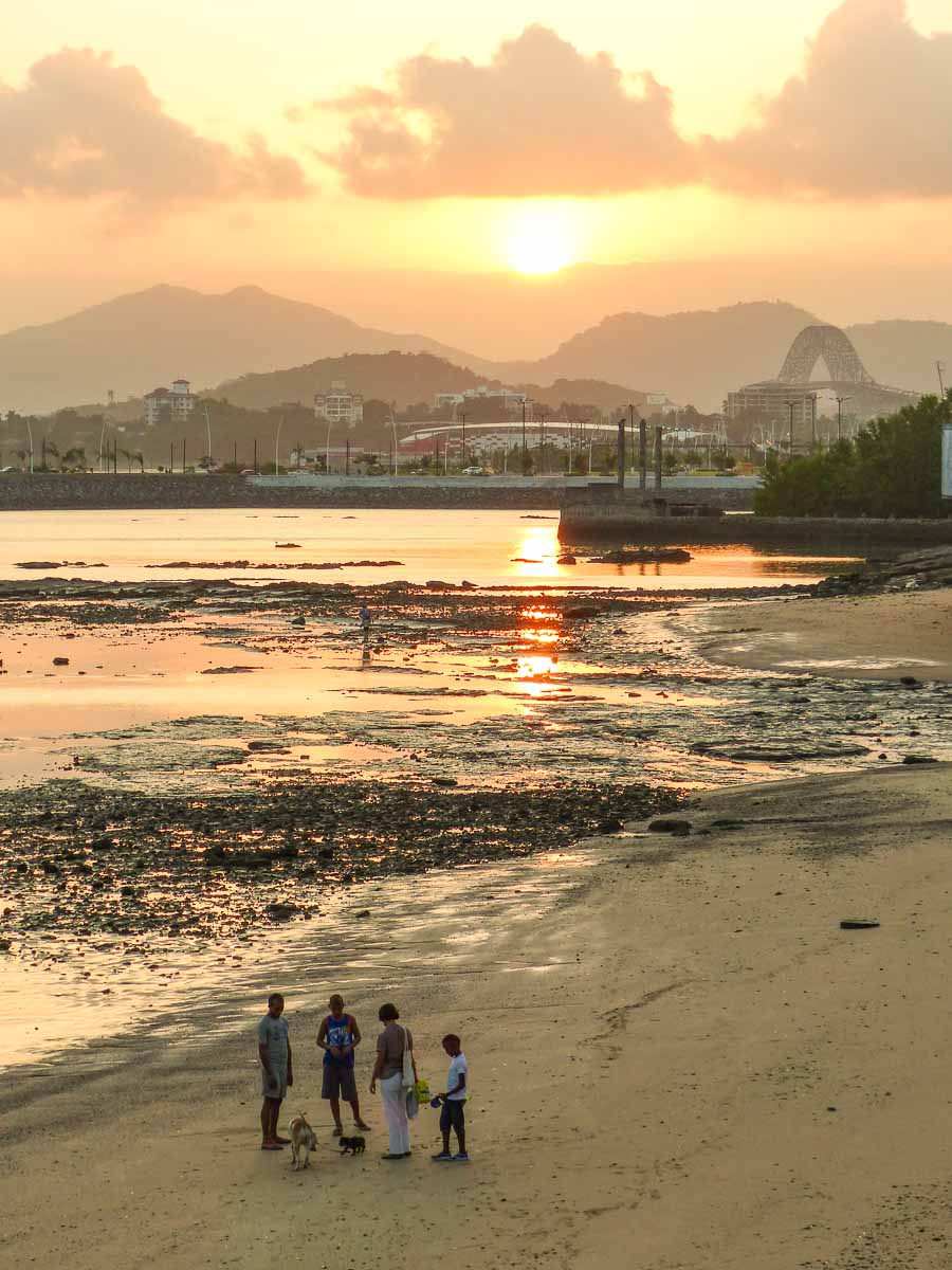 Eine Familie geht bei Sonnenuntergang am Strand in Panama City spazieren. Im Hintergrund sieht man ein Schiff, die Puente de las Americas über den Panamakanal und Berge.