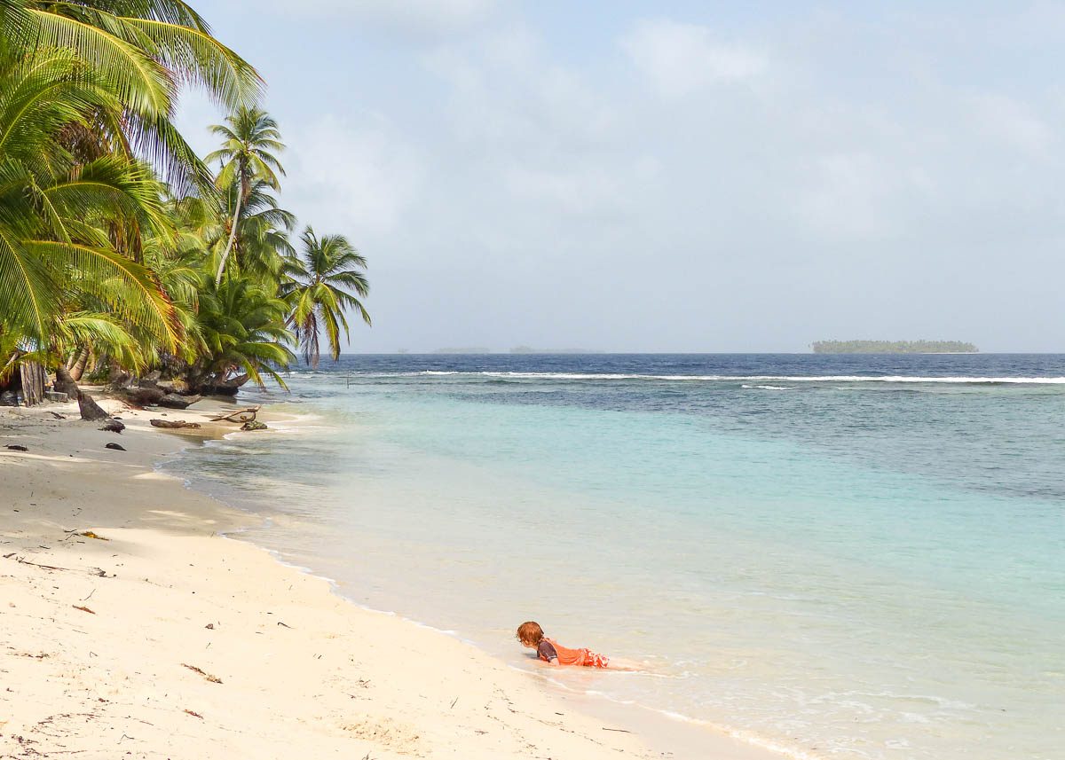 Ein Kind liegt halb im türkisblauen Wasser halb am weißen Strand mit Palmen und tropischer Vegetation. Im Hintergrund sieht man eine karibische San Blas Insel.