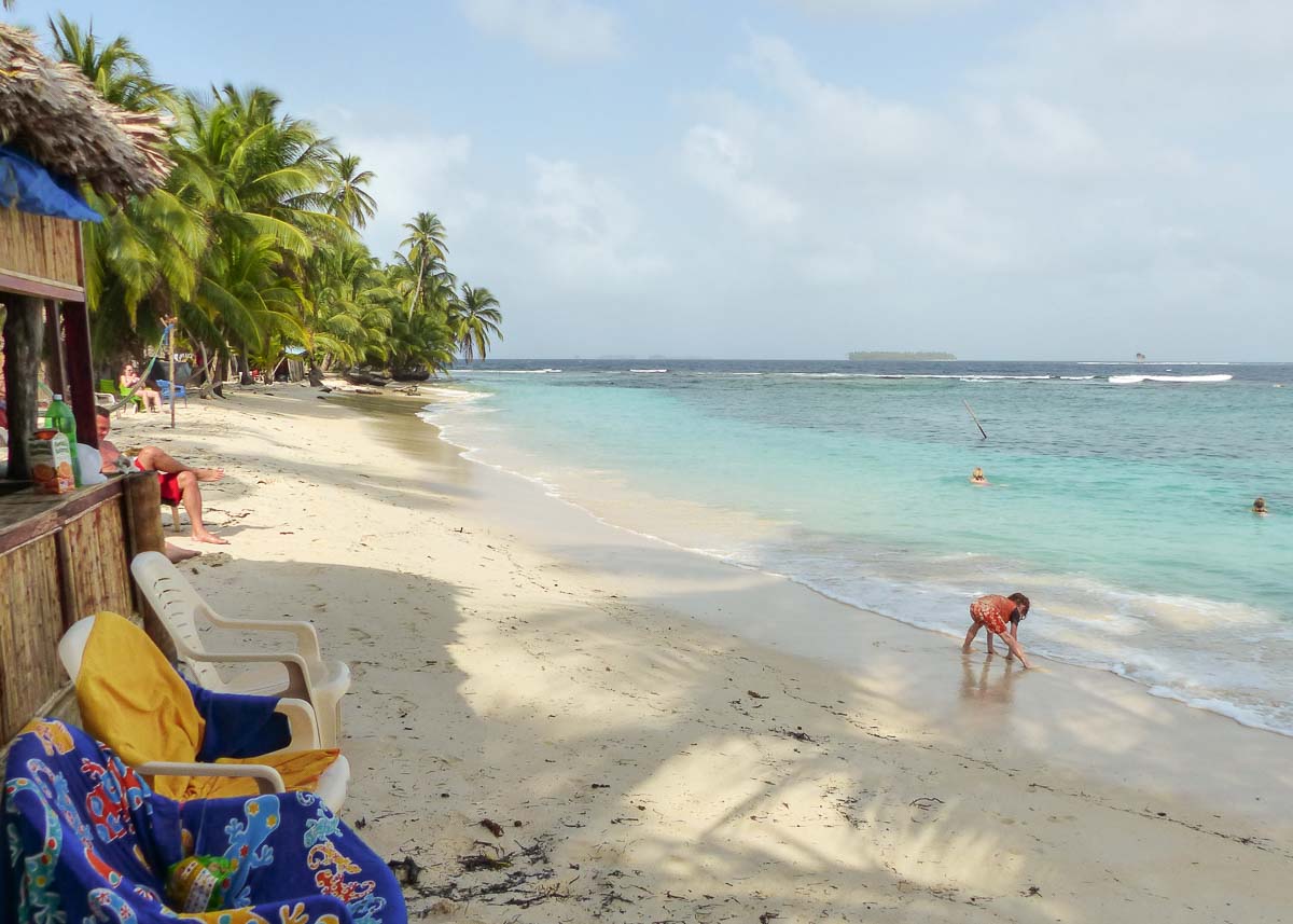 Ein Kind spielt im türkisblauen Wasser an einem weißen Strand auf den San Blas Inseln in Panama. Im Vordergrund stehen Strandstühle mit Handtüchern. Im Hintergrund sieht man eine andere Insel. Der Strand ist gesäumt von Palmen und menschenleer.