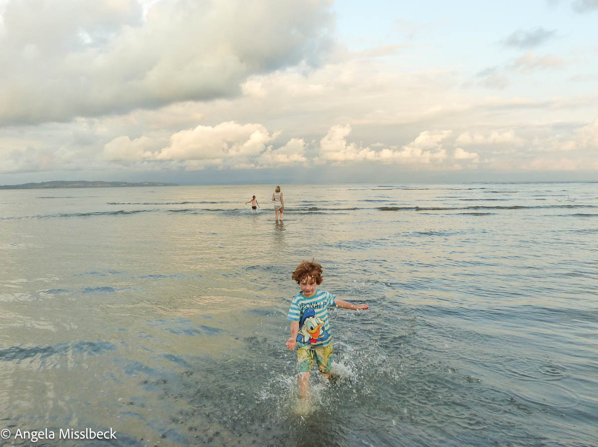 Ein kleines Kind spielt im knietiefen Wasser, im Hintrgrund läuft eine Mutter mit Kind durch flaches Wasser an einem Strand bei Abendstimmung.
