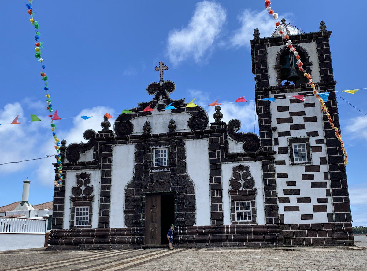 Die weiße Fassade der eher flachen Kirche Santo Espiritu mit gedrungenem quadratischen Glockenturm auf der Insel Santa Maria, Azoren, ist unterbrochen von dunklem Vulkangestein