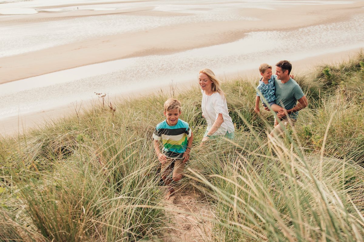 Eine Familie mit zwei Kindern spaziert durch Dünen. Der Vater trägt ein Kind.