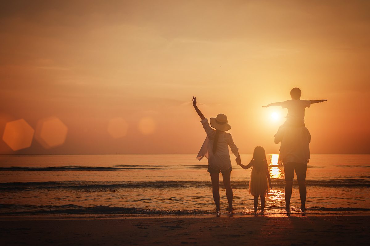 Eine Familie steht mit ausgebreiteten Armen im Sonnenuntergang am Meer.