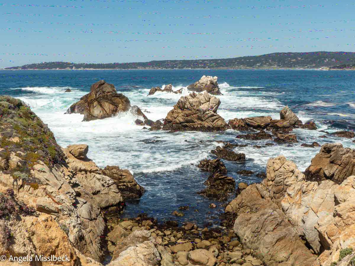 Felsige Küste mit Wellen, die gegen große Felsen schlagen, unter einem klaren, blauen Himmel, typisch für den Westen der USA. Das Meer erstreckt sich bis zum Horizont, im Hintergrund ist die zerklüftete Küste des Point Lobos Naturparks Kalifornien zu sehen. Zwischen den Felsen im Vordergrund wächst spärliche Vegetation.