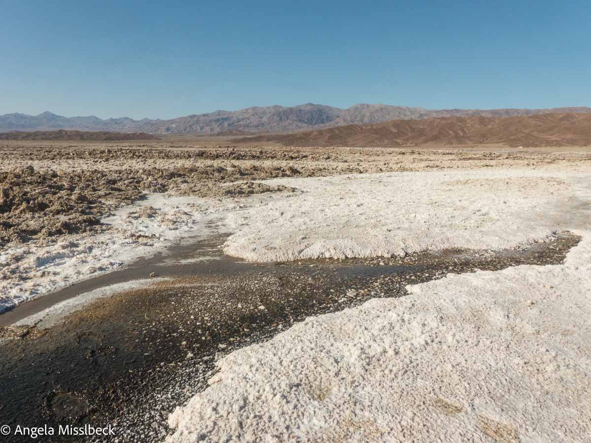 Ausgedehnte Salzebenen erstrecken sich über den Death Valley Nationalpark im Westen der USA. Unter einem klaren, blauen Himmel bilden ferne Bergketten die Kulisse für ein trockenes Gelände, in dem sich weiße Salzkruste mit Flecken karger Erde vermischt.