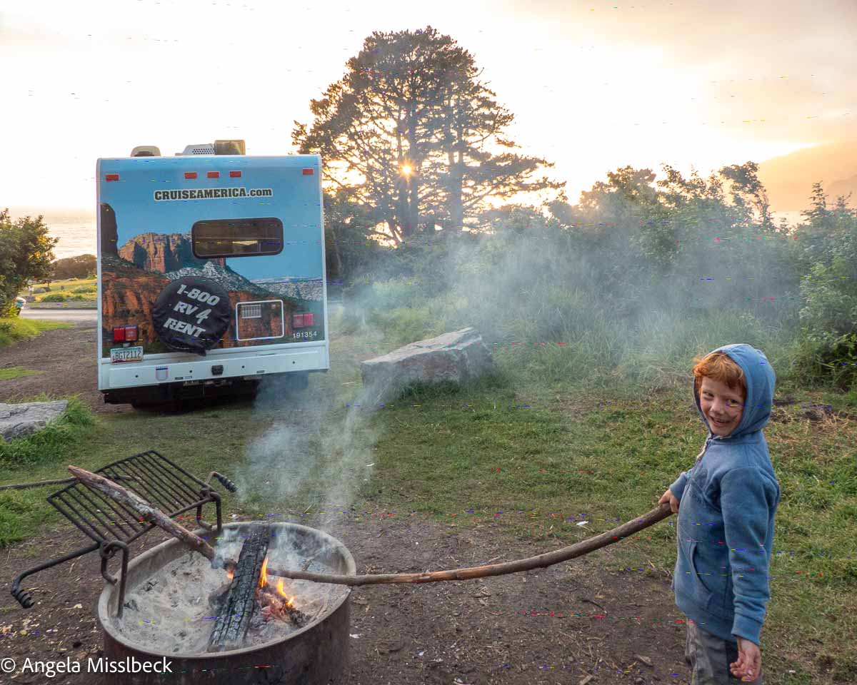 Ein Kind in einem blauen Kapuzenpullover lächelt und hält einen langen Stock über ein Lagerfeuer. Ein gemietetes Wohnmobil steht in der Nähe, umgeben von der ruhigen Schönheit der Küstenparks im Westen der USA. Bäume zeichnen sich vor einem leuchtenden Sonnenuntergang ab, während sanft Rauch vom Feuer aufsteigt.