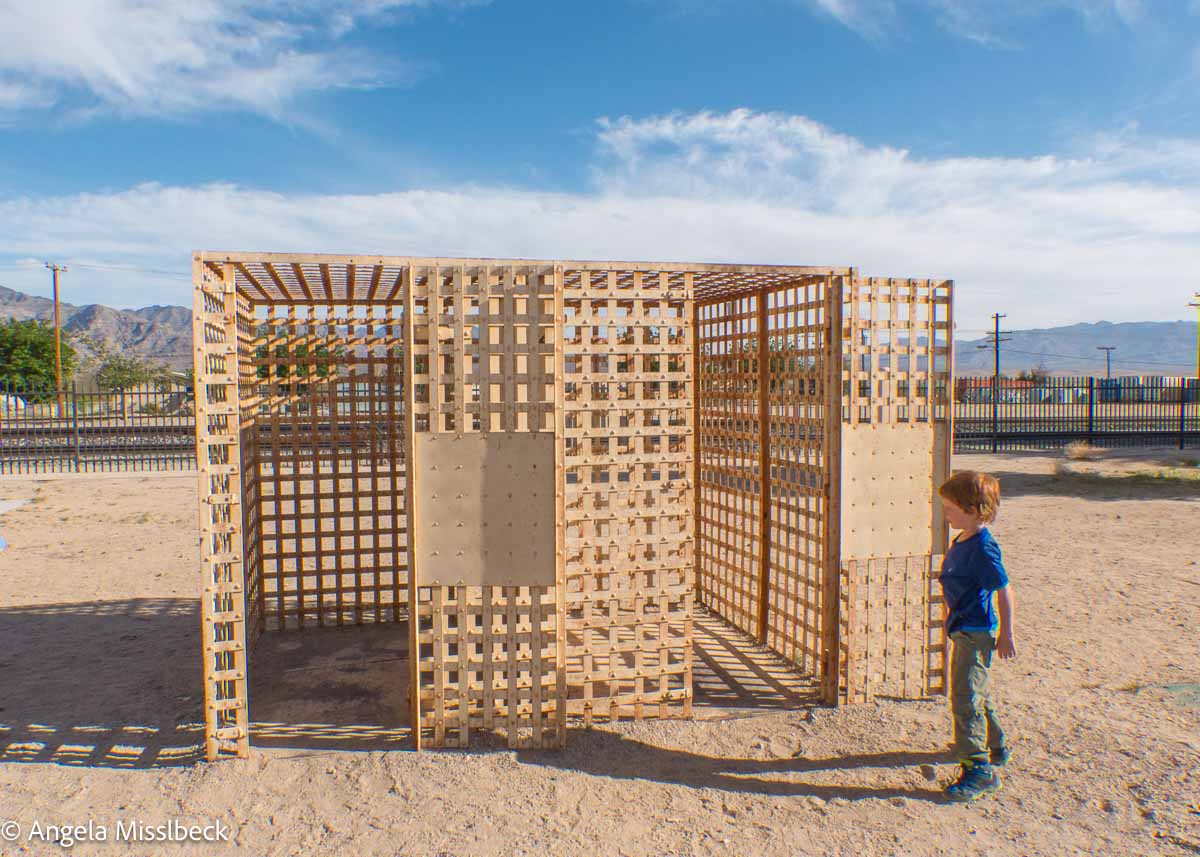 Ein kleines Kind betrachtet eine große, offene, würfelartige Struktur aus Metallgitter , die auf sandigem Boden steht, mit klarem blauen Himmel und Bergen in der Ferne, was an die weiten Landschaften der Nationalparks der USA erinnert.