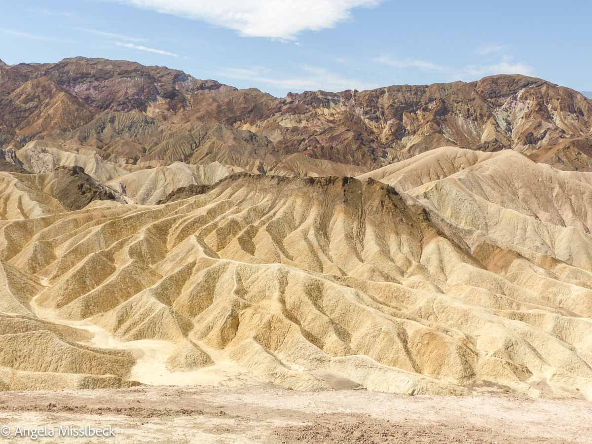 Ein Panoramablick auf schroffe, erodierte Hügel mit geschichteten Erdtönen unter einem teilweise bewölkten Himmel zeigt die einzigartigen geologischen Formationen und Farben der Wüstenlandschaft am Zabriskie Point im Death Valley Nationalpark der USA.