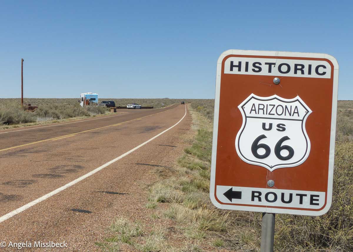 Eine Wüstenstraße mit einem Schild der historischen Arizona Route 66 im Vordergrund erstreckt sich unter einem klaren blauen Himmel. Der berühmte Highway, umgeben von der weitläufigen Schönheit der US-Nationalparks, erstreckt sich in die Ferne, gesprenkelt mit ein paar vereinzelten Fahrzeugen.