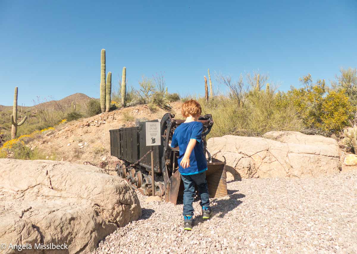 Ein kleines Kind in blauem Hemd und Jeans steht neben einem alten Bergbauwagen auf einem Wüstenpfad und verkörpert den Geist des amerikanischen Westens. Kakteen, Sträucher und ein strahlend blauer Himmel rahmen die Szenerie vor felsigem Gelände in dieser malerischen Natur- und Nationalparkkulisse ein.