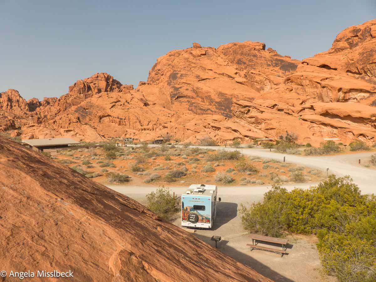 Ein Wohnmobil parkt in einer malerischen Wüstenlandschaft im Westen der USA, umgeben von roten Sandsteinformationen und spärlicher Vegetation unter einem klaren blauen Himmel. Ein Picknicktisch lädt zum Entspannen ein, während sich im Hintergrund majestätisch schroffe Klippen erheben.