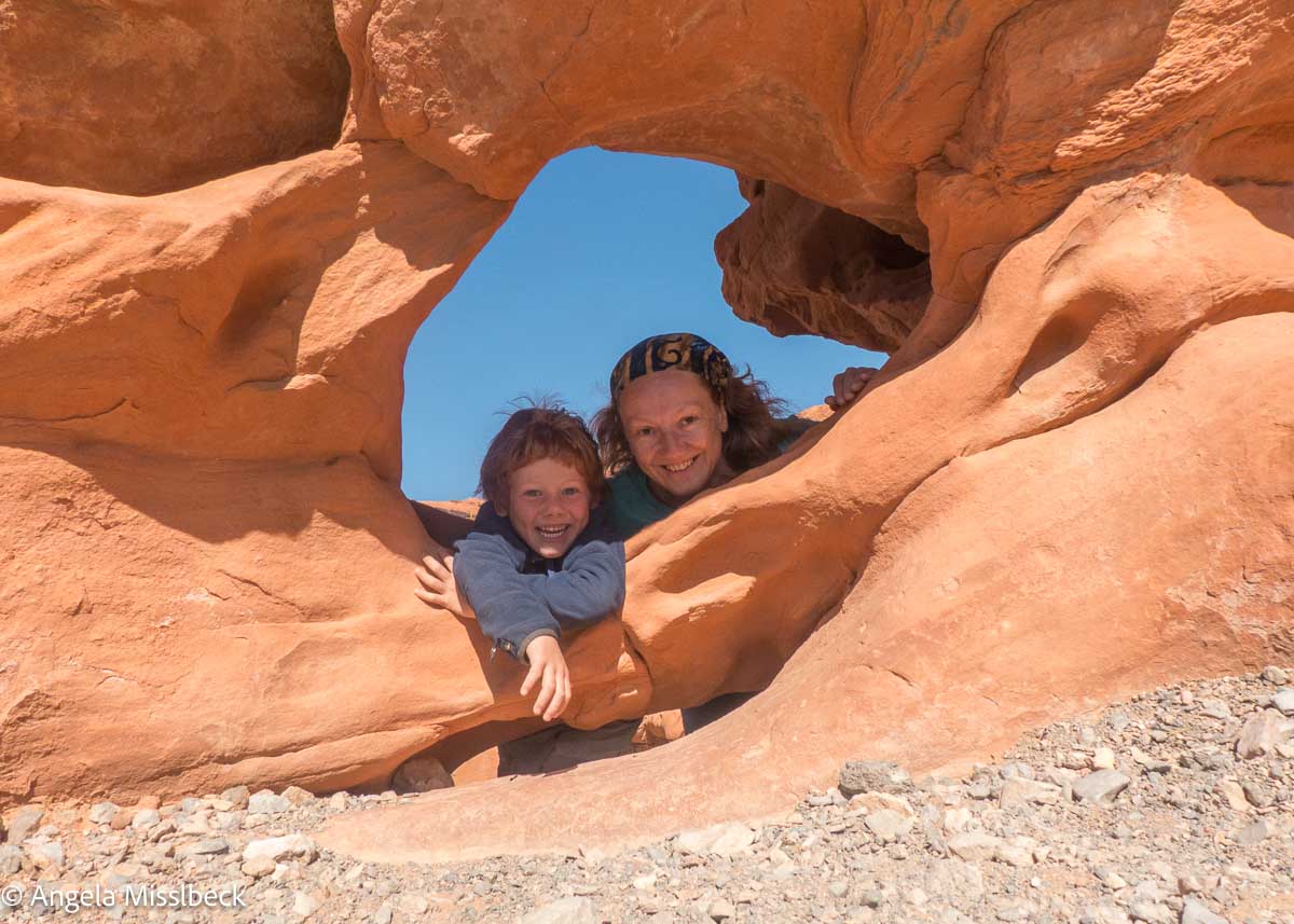 Eine Mutter und ihr Kind lächeln und spähen durch ein Felsenfenster im Valley of Fire State Park (Nevada). Die orangefarbenen, strukturierten Felsen heben sich vom klaren blauen Himmel ab, während Kieselsteine und kleine Steine den Boden bedecken.