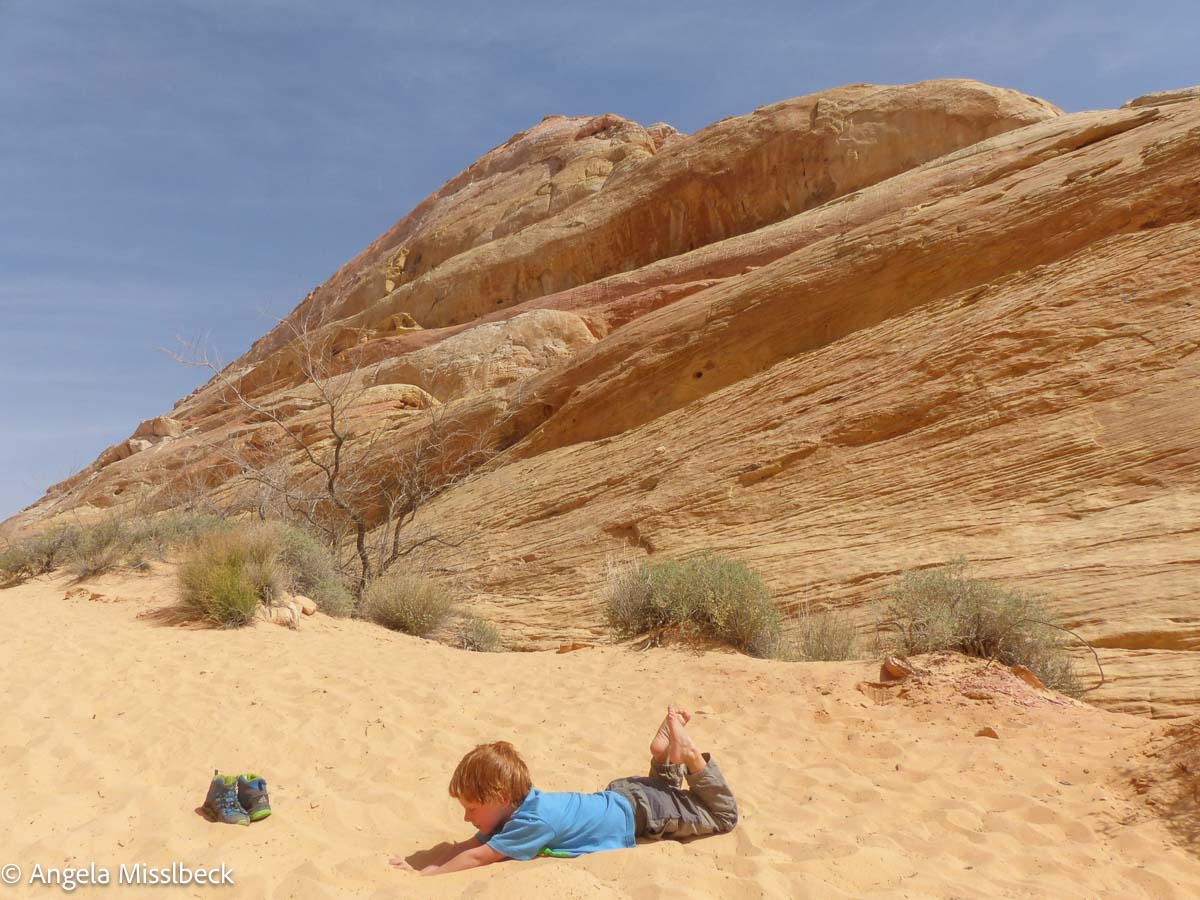 Ein Kind liegt bäuchlings im Sand und blickt auf einen großen, felsigen Hügel in einem der Naturparks der USA. Der Himmel ist klar, die Vegetation spärlich. Das Kind trägt ein blaues Hemd und eine blaue Hose, daneben stehen Schuhe.