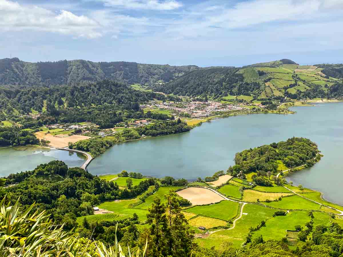 Ein malerischer Blick auf São Miguel mit Kindern: üppig grüne Landschaft mit einem gewundenen See, umgeben von Feldern, Bäumen und Hügeln unter einem blauen Himmel mit vereinzelten Wolken. In der Ferne ist entlang der Küste eine kleine Stadt zu sehen.