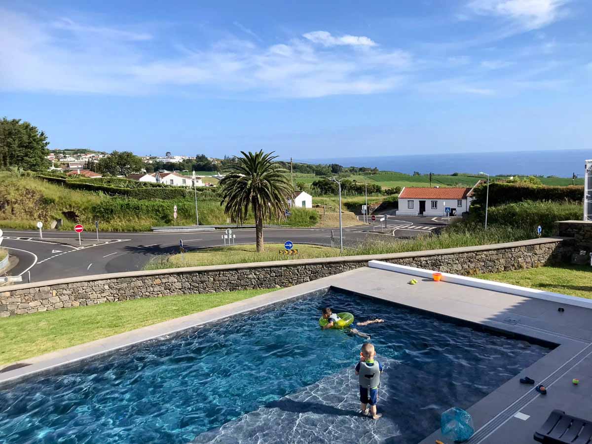 Zwei Kinder spielen in einem Swimmingpool auf São Miguel mit Blick auf eine malerische Landschaft mit grünem Gras, Palmen, gewundenen Straßen, weißen Häusern und dem Meer unter einem klaren blauen Himmel.