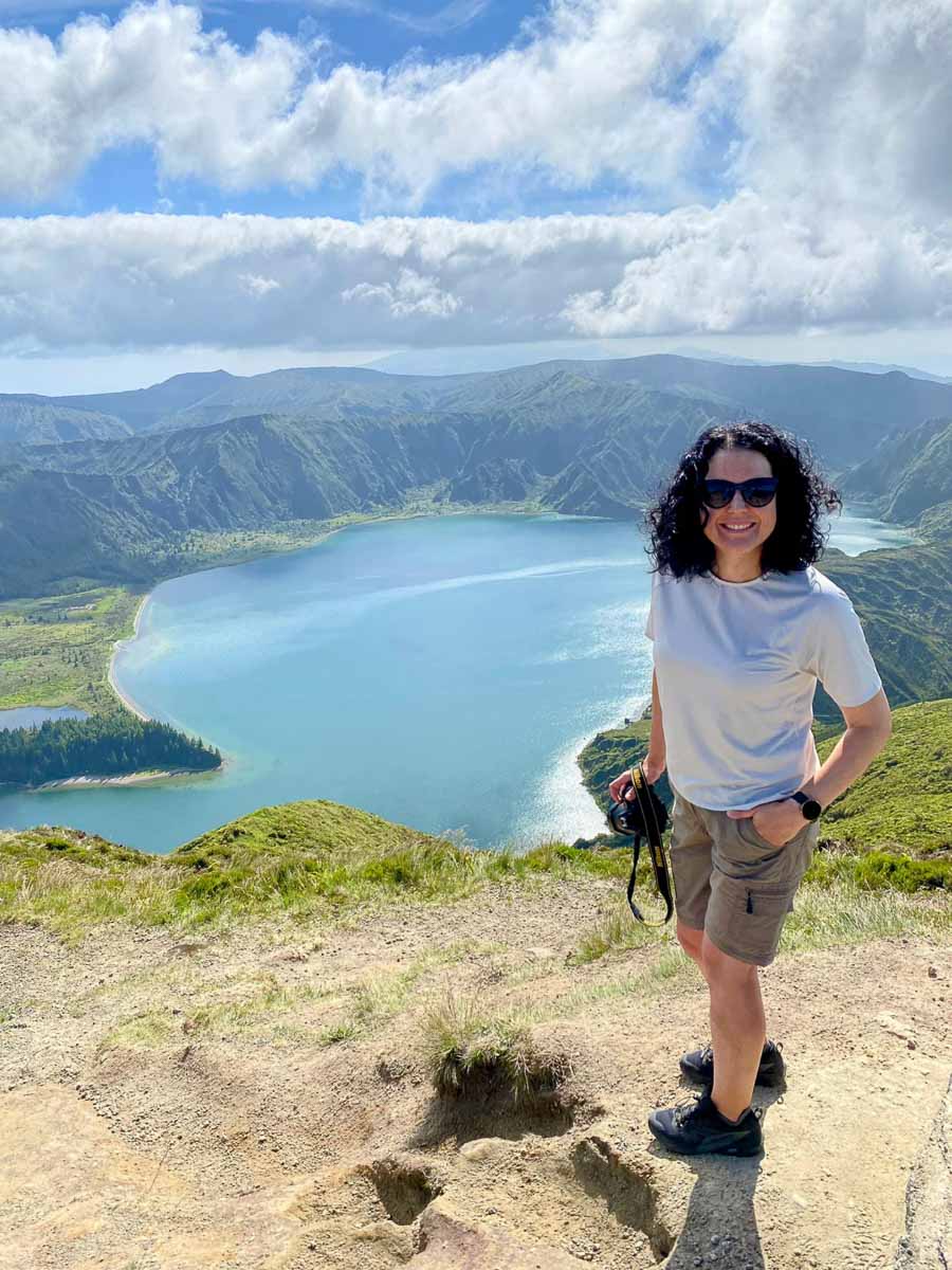 Eine Frau mit Sonnenbrille steht lächelnd und mit einer Kamera in der Hand auf einem grasbewachsenen Hügel und genießt die atemberaubende Aussicht auf São Miguel mit Kindern – der große blaue See Lagoa do Fogo und die grünen Berge erstrecken sich hinter ihr unter einem teilweise bewölkten Himmel.