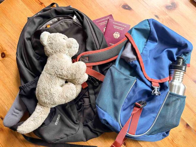 Zwei Rucksäcke, eine Plüschkatze für Babys, zwei Reisepässe und eine Wasserflasche aus Metall liegen auf einer hölzernen Unterlage - alles, was man für das Handgepäck mit Kindern braucht und mit Checklisten für eine bevorstehende Reise zusammenstellt.