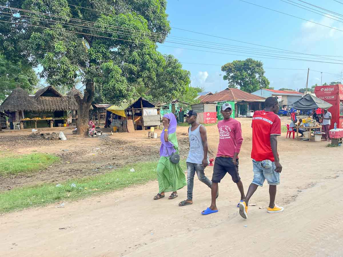 Vier Personen gehen in Kenia eine unbefestigte Straße in einem ländlichen Dorf entlang, vorbei an Bäumen, Marktständen und strohgedeckten Hütten. Kleine Geschäfte und verstreute Stühle säumen den Straßenrand und fangen den Geist des täglichen Lebens in Kenia ein.