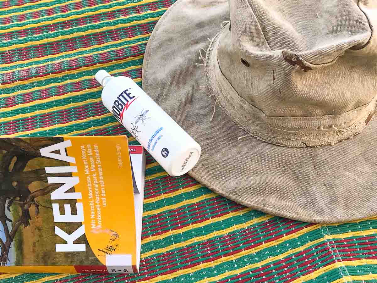 Ein abgenutzter beiger Hut, Insektenschutzmittel und ein KENIA-Reiseführer liegen auf einer bunten gewebten Matte, die ideal für einen Familienurlaub in Kenia ist.