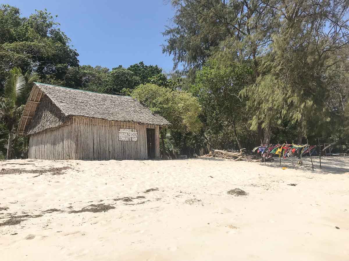 Eine Holzhütte mit Strohdach steht an einem von Bäumen umgebenen Sandstrand. Rechts hängen bunte Kleider auf einer Leine im Schatten - eine friedliche Szene, wie sie für eine Kenia-Reise typisch ist.