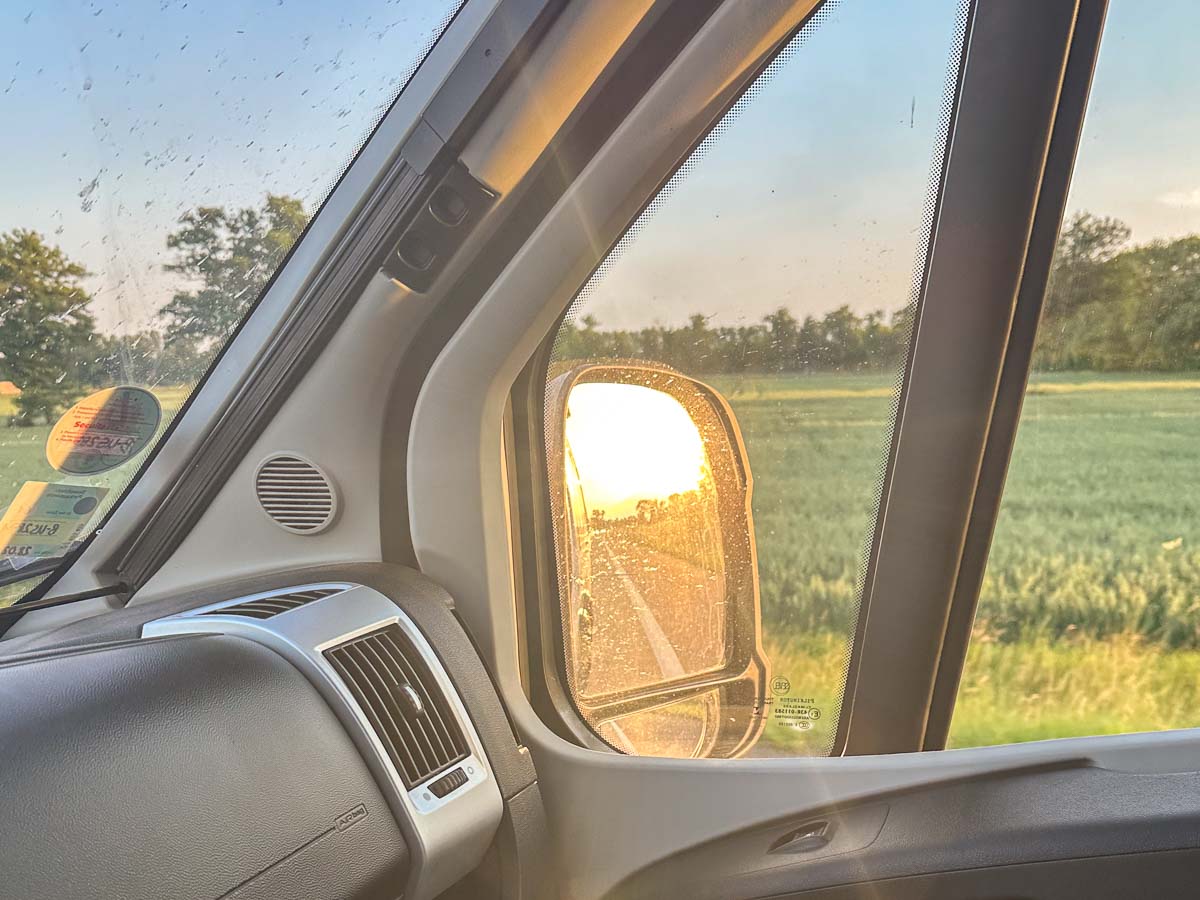 Blick aus dem Inneren eines Fahrzeugs während eines Roadtrips mit Kindern, mit der sich im Seitenspiegel reflektierenden Sonne, grünen Feldern und Bäumen vor dem Fenster und dem Armaturenbrett und Fensterrahmen im Vordergrund.