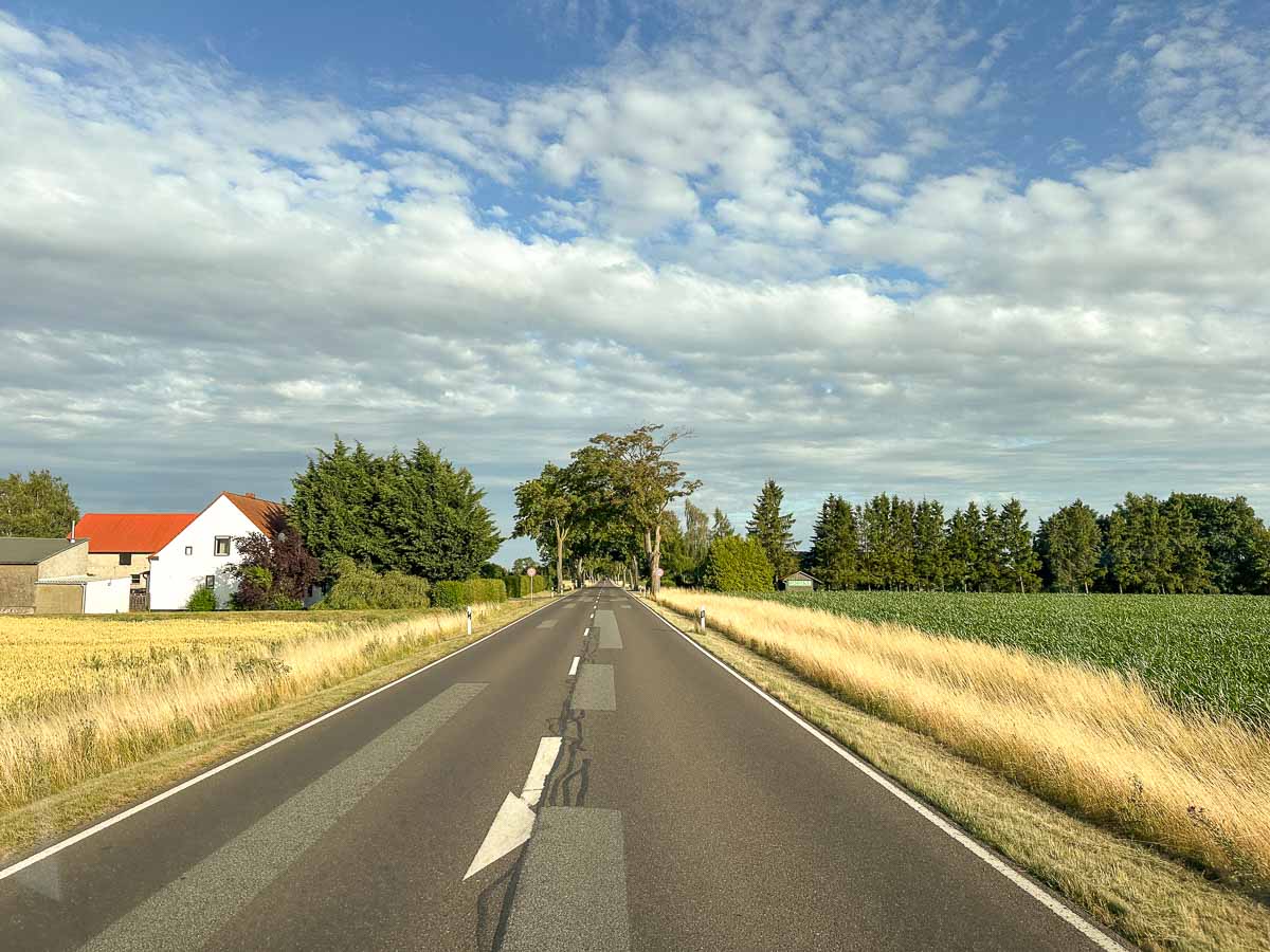 Eine leere zweispurige Straße führt durch Ackerland mit Feldern auf beiden Seiten, perfekt für einen Roadtrip, ein weißes Haus mit rotem Dach auf der linken Seite, Bäume am Horizont und ein teilweise bewölkter blauer Himmel darüber.