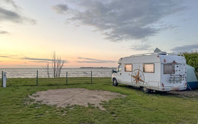 Ein weißes Wohnmobil steht bei Sonnenuntergang auf einer Wiese neben einem Metallzaun auf einem Ostsee Campingplatz am Strand und blickt auf das ruhige Meer.