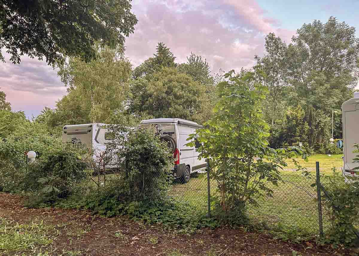 Zwei weiße Wohnmobile stehen auf einer Wiese, die von üppigem Grün und Bäumen umgeben ist. Ein Metallzaun und  Büsche sind im Vordergrund unter einem teilweise bewölkten Abendhimmel zu sehen.