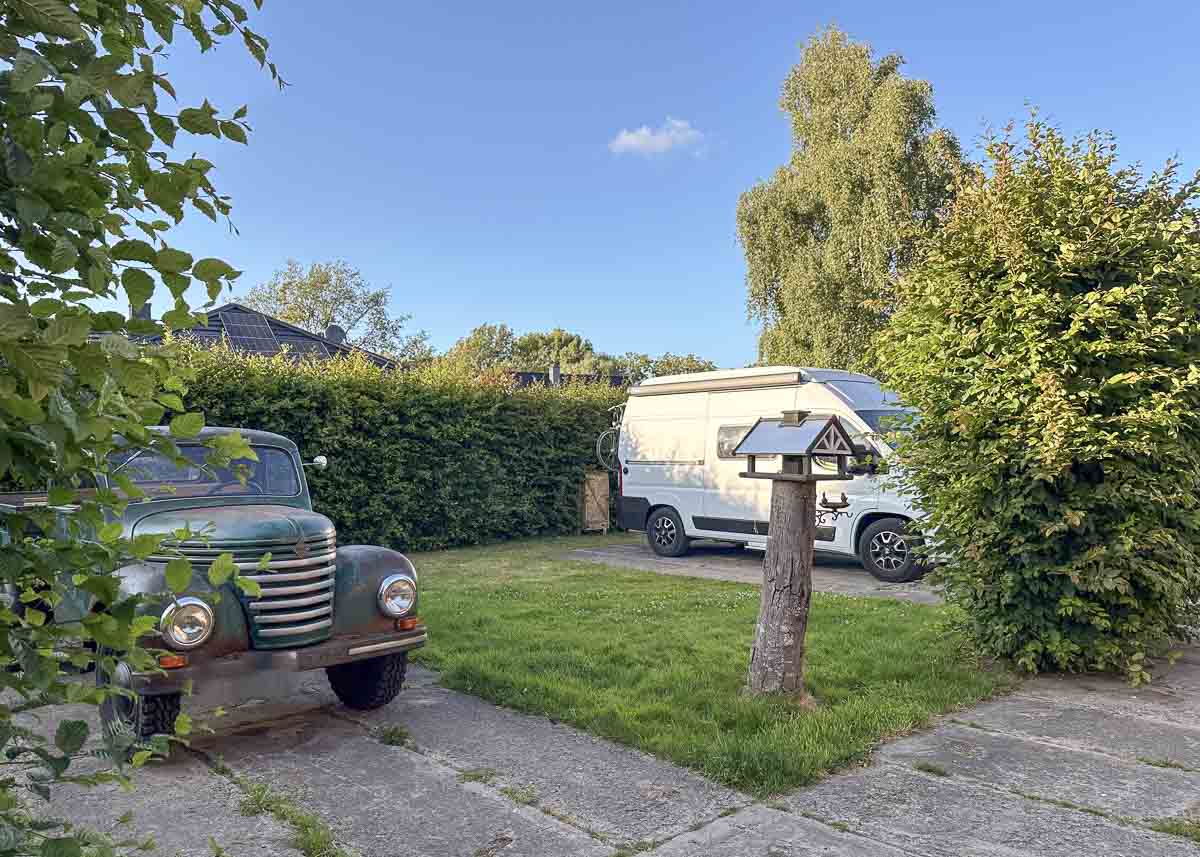Ein grüner Oldtimer und ein weißes Wohnmobil stehen auf einer von Hecken gesäumten Wiese auf einem charmanten Ostsee-Wohnmobilstellplatz, mit einem hölzernen Vogelhaus auf einem Pfosten in der Mitte und Bäumen unter dem klaren blauen Himmel.
