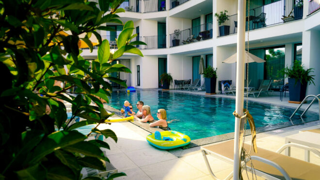 Mehrere Kinder spielen auf aufblasbaren Spielzeugen in einem familienfreundlichen Hotelpool, umgeben von modernen Balkonen und Liegestühlen, mit grünen Pflanzen im Vordergrund und Sonnenlicht, das die Szene hervorhebt.