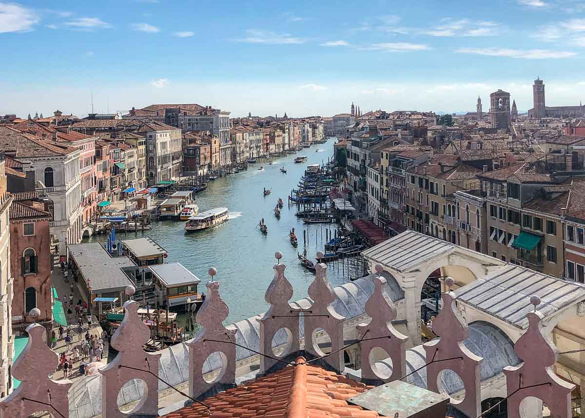 EUROPA-Venedig-0087 Ein malerischer Blick auf den Canal Grande in Venedig mit Booten, historischen Gebäuden zu beiden Seiten und kunstvoll verzierten Dachbalustraden im Vordergrund unter strahlend blauem Himmel.