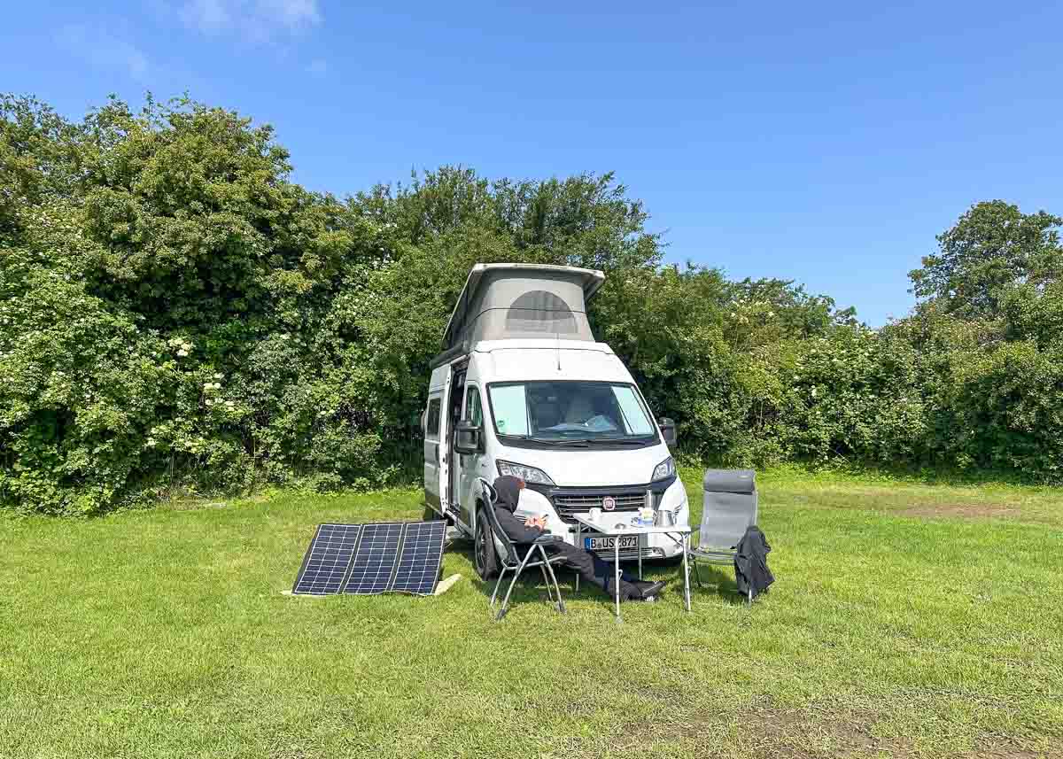 Ein weißer Wohnmobil mit ausgefahrenem Dach steht auf einer Wiese in der Nähe von Büschen. Ideal, um Fehmarn mit Kindern zu erkunden: Unter strahlend blauem Himmel stehen draußen Sonnenkollektoren, zwei Stühle und ein kleiner Tisch bereit, an dem ein Teenager in dunkler Kleidung sitzt.