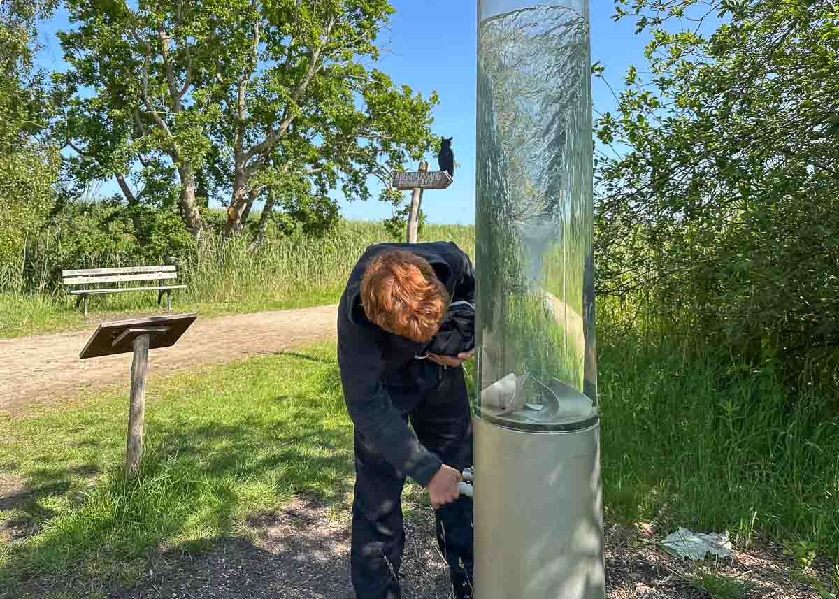 Ein Teenager mit rötlichem Haar bedient eine Handpumpe, die an einem hohen Wasserzylinder befestigt ist, im Naturschutzgebiet Wallnau auf Fehmarn mit Rasenfläche, Bäumen, einer Bank und einem Feldweg 
