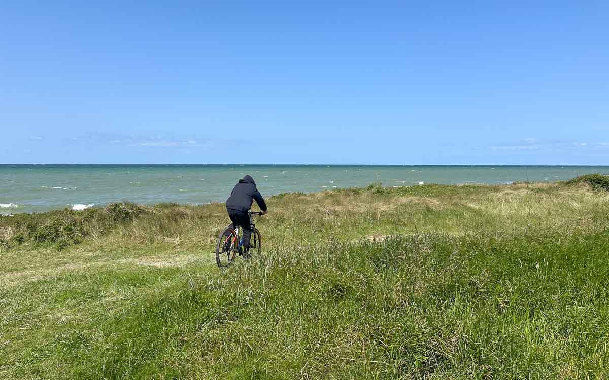 Ein Teenager in einem dunklen Kapuzenpulli fährt unter strahlend blauem Himmel auf einem grasbewachsenen Weg auf Fehmarn mit dem Fahrrad in Richtung Meer. In der Ferne schimmern die Wellen.
