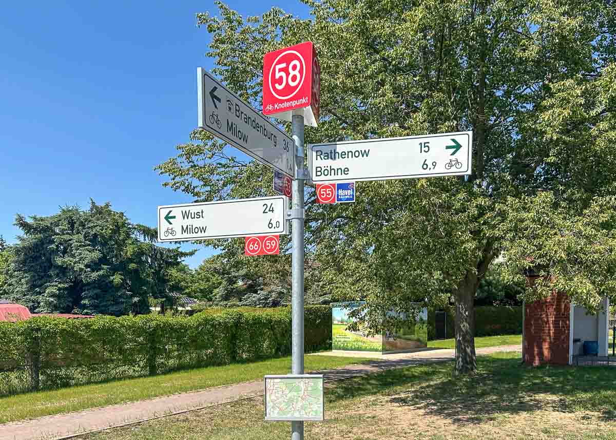 Ein Wegweiser aus Metall mit Radwegnummern und Zielorten weist in Richtung Rathenow, Böhne, Wust, Milow und Brandenburg im Havelland. Im Hintergrund sind an einem sonnigen Tag ein Baum, Häuser und ein Weg zu sehen.